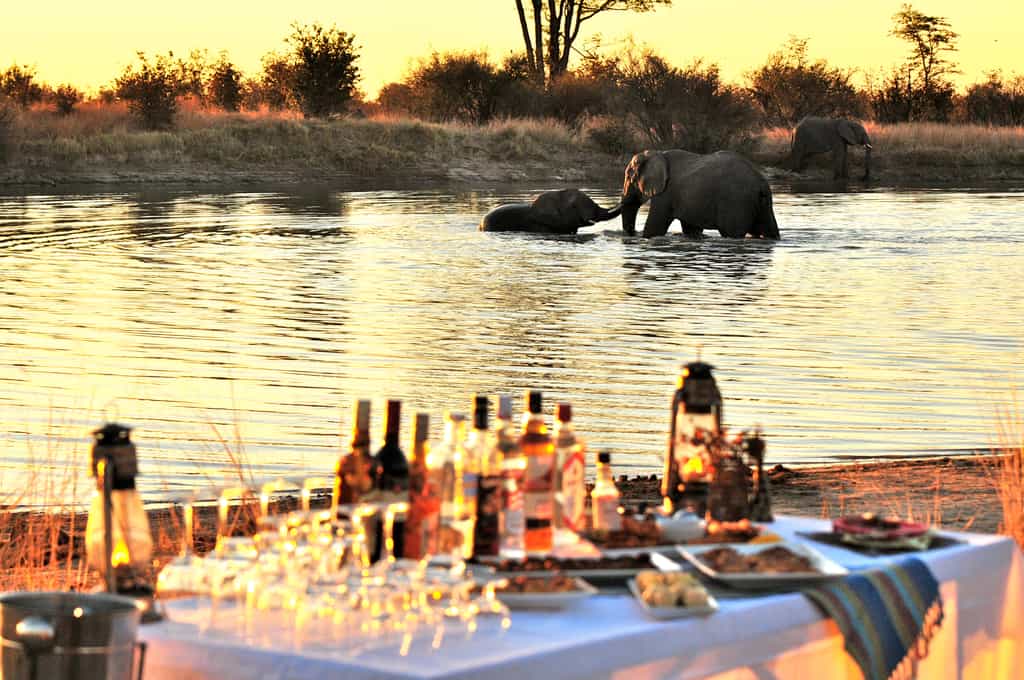 Elephants splashing in the pan while guests enjoy sundowners at sunset
